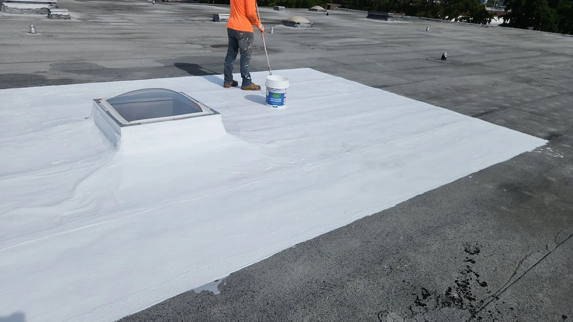 Roofing technician applying white silicone coating around a skylight on a flat roof